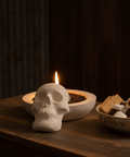 Skull-shaped concrete candle holder with a lit candle on a wooden table, with a bowl of S'mores.