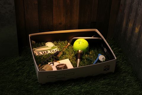 Golf-themed items including a golf ball, club, and hat in a box on grass with a wooden background.
