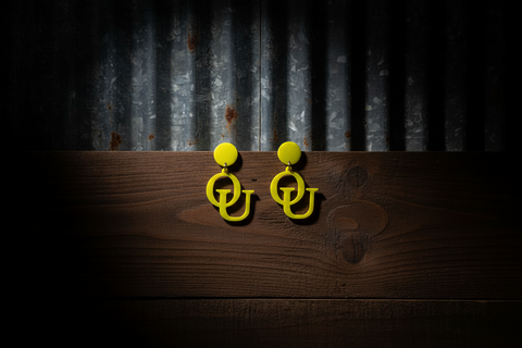 Yellow earrings with 'OU' design on a wooden surface and corrugated metal background