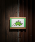 Hand-drawn illustration of a green dinosaur with party hats on a wooden board against a dark wooden background.
